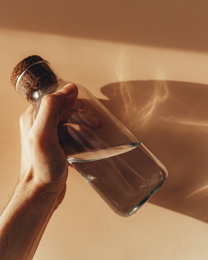 Crop anonymous male holding transparent glass bottle of water in hand against beige wall with shades during sunny day